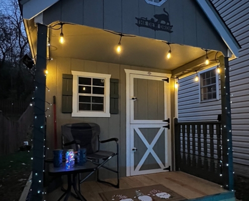 A cozy outdoor shed illuminated by string lights at twilight, with a chair and decor on a small porch
